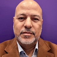 Man with brown skin and a goatee smiles at the camera. He is wearing a brown coat with a blue shirt. 