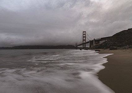 Water flowing onto beach on a cloudy day with Golden Gate Bridge in background