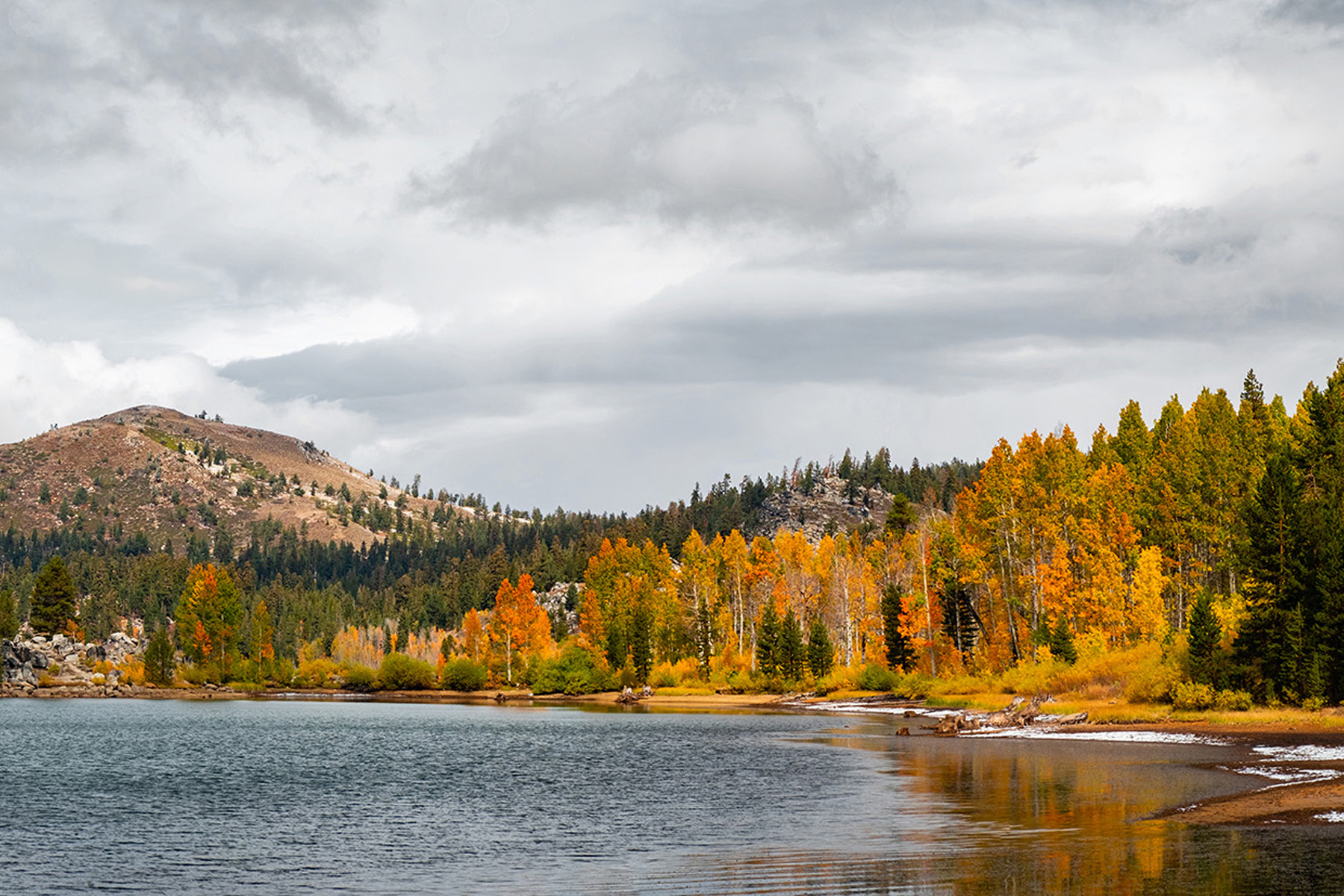 Photo shows a lake and in the background are autumnal-colored tress and a small mountain. 