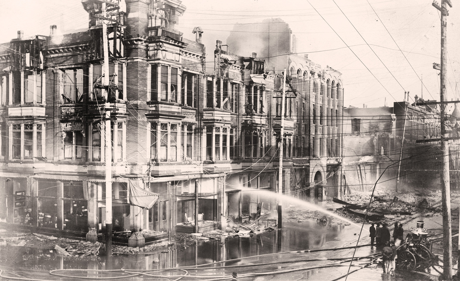 epia-toned image shows a multistory burnt-out building being sprayed by firemen. 