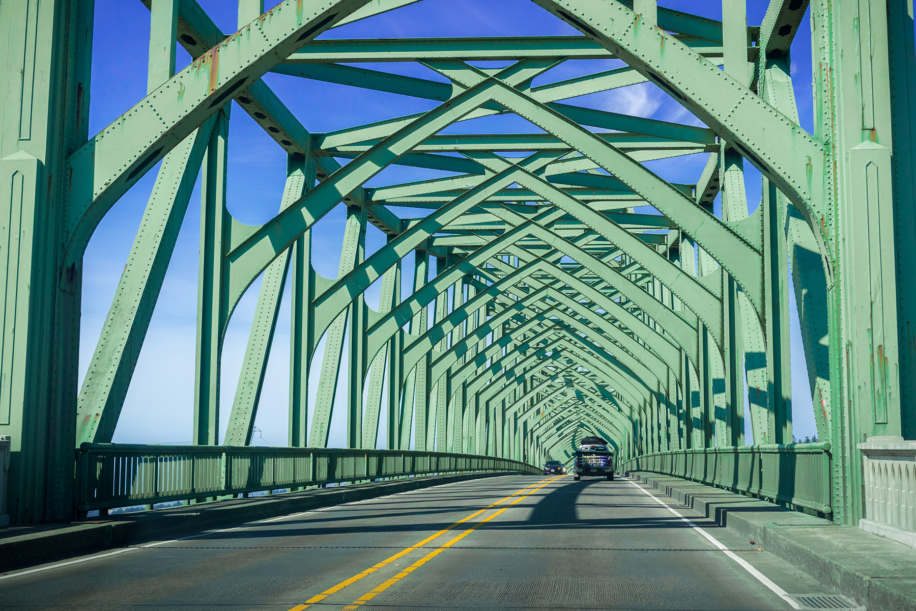 Cross-bracing painted green over a roadway is shown. A car travels along the roadway. 