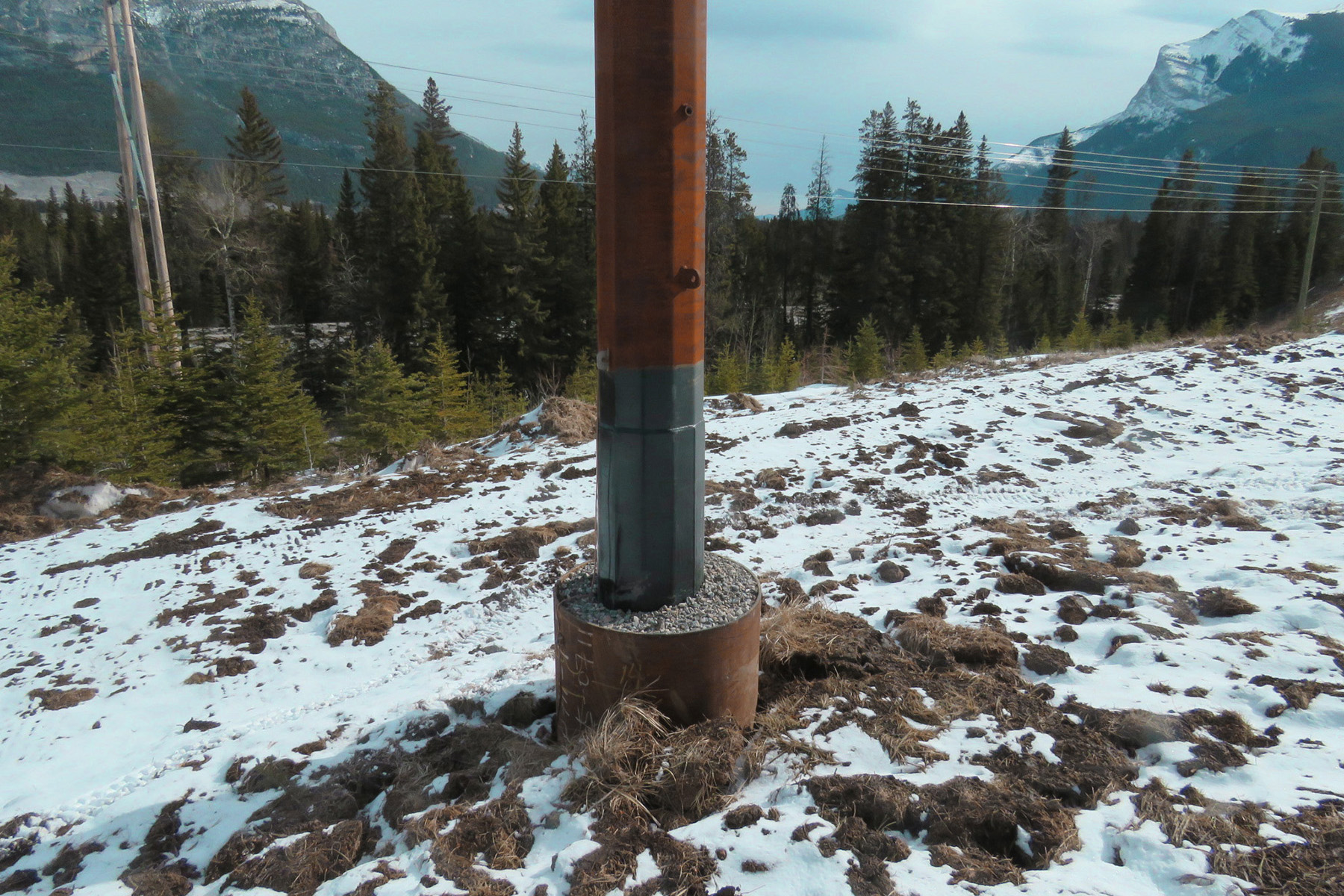 A wooden pole in a backfilled bucket foundation stands in a snowy field. 