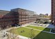Three multistory buildings surround a large open outdoor space with sidewalks, a grass lawn, and trees. People walk on the sidewalks. 