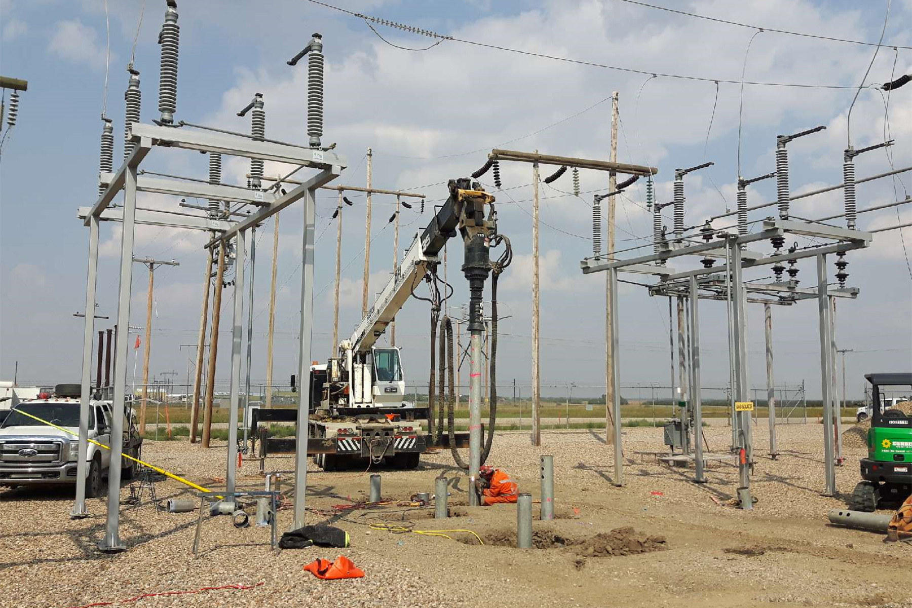 A piece of construction equipment drives steel helical piles into the ground within a constrained area with electrical wires and equipment overhead. 