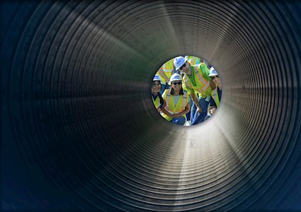 Four students wearing yellow safety vests, black sunglasses, and white hard hats smile into a ridged tunnel.