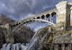 Water cascades over stone dam beneath arched bridge and cloudy sky