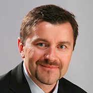 A man with a mustache and brown hair, wearing a white collared shirt and black jacket, smiles at the camera. 